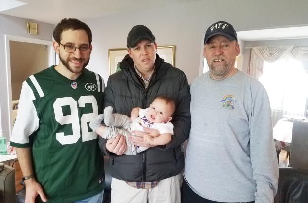 Three men stand in a living room smiling for a photo. One is holding a baby.