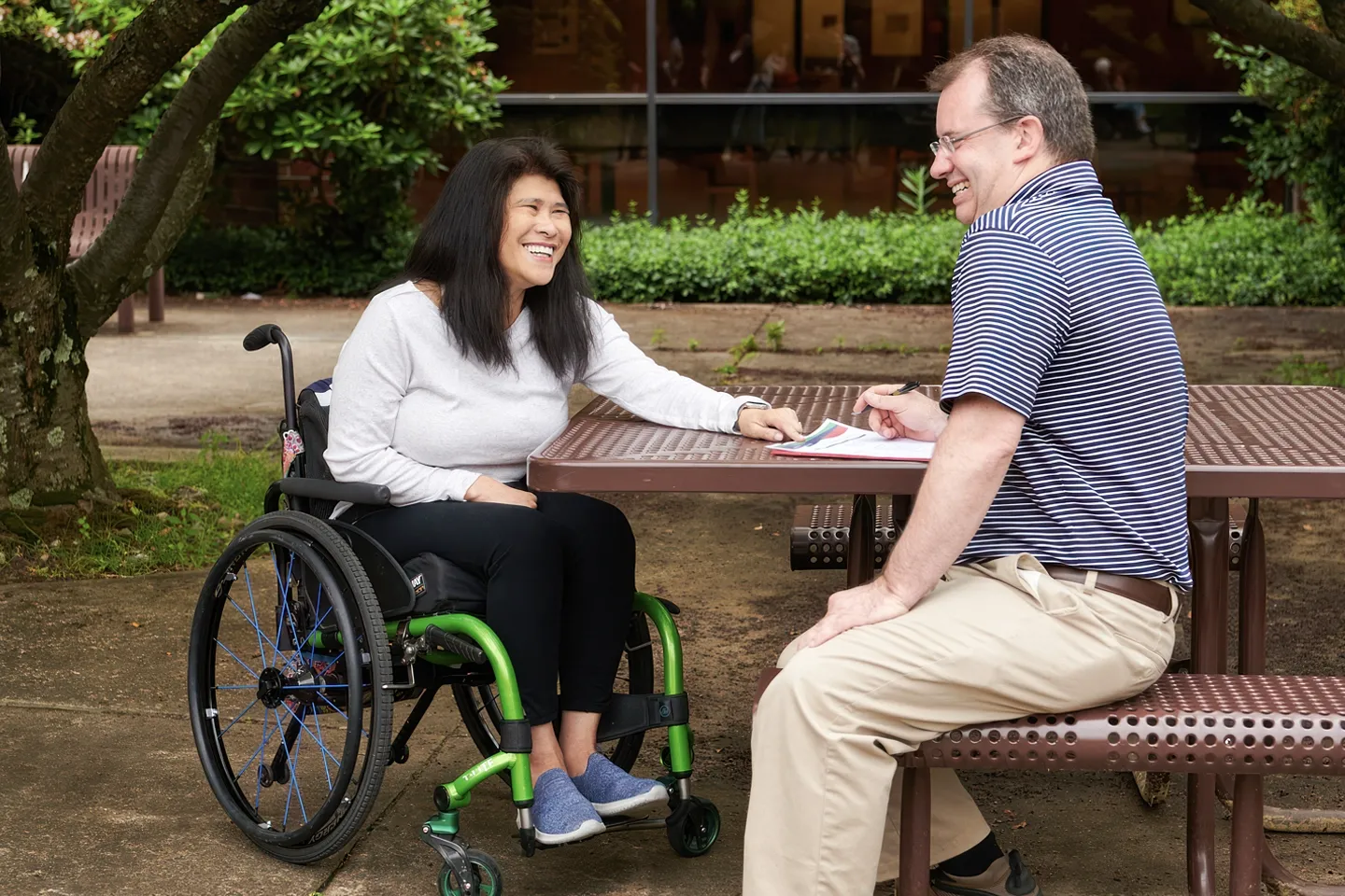 A woman is sitting in a wheelchair next to her male personal care assistant at an outside table surrounded by greenery. Both are smiling broadly.