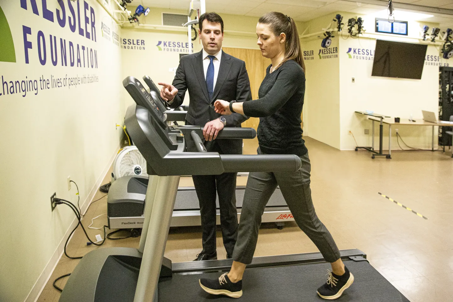 Woman on treadmill exercises while male researcher in business suit observes standing next to the machine.