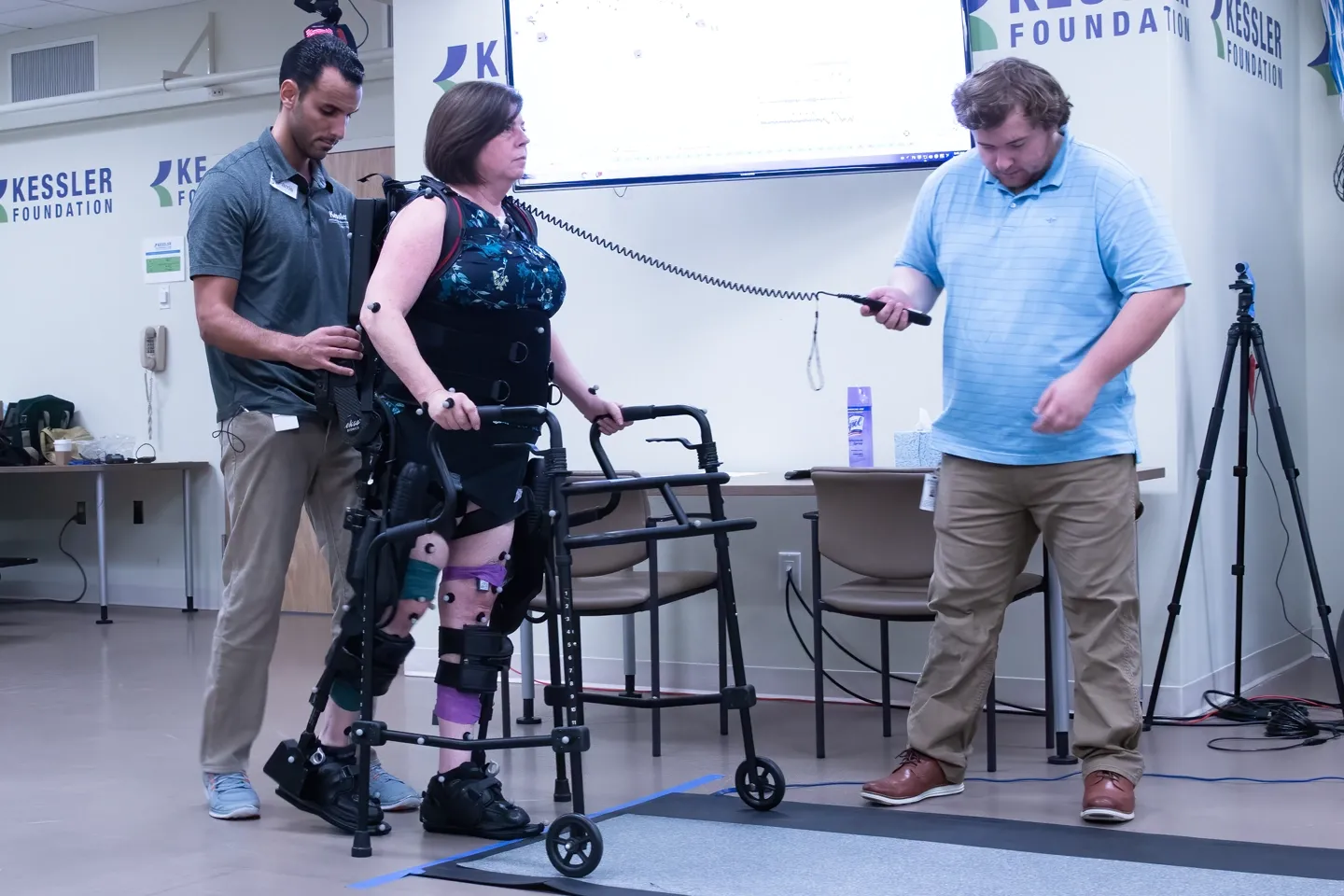 Woman walks wearing an exoskeleton with the help of two male research assistants, one standing behind her while the other stands a few feet away adjusting the machine.