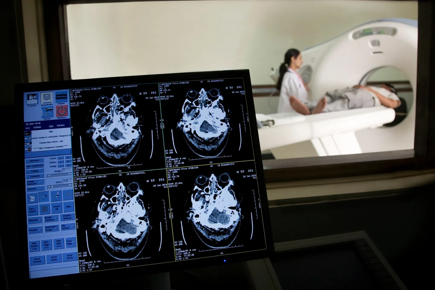 Image of MRI results are shown on a computer screen in foreground; technician with someone in an MRI scanner in the background.