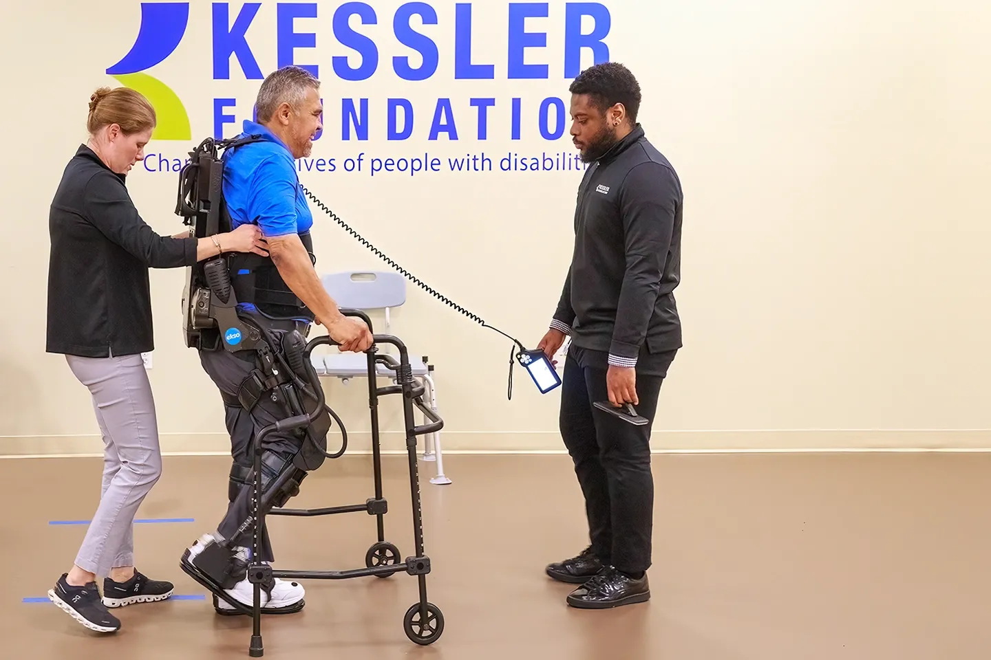 Christina Dandola, PhD, physical therapist at the Center for Mobility and Rehabilitation Engineering Research steadies a male stroke patient exercising in an EksoGT exoskeleton from behind, with Corey Greene, research assistant, looking on in front.