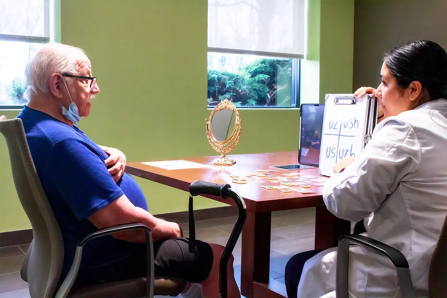 A female clinician holding a board with various letters to help a man sitting across the table with a stroke-induced reading impairment.