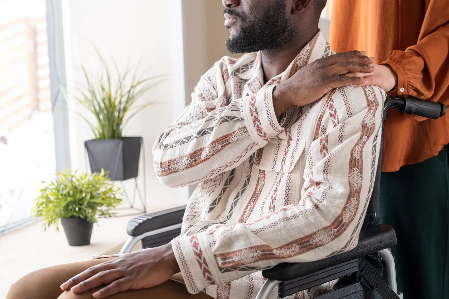 Picture of black man sitting in a wheelchair being comforted by a white individual from behind.