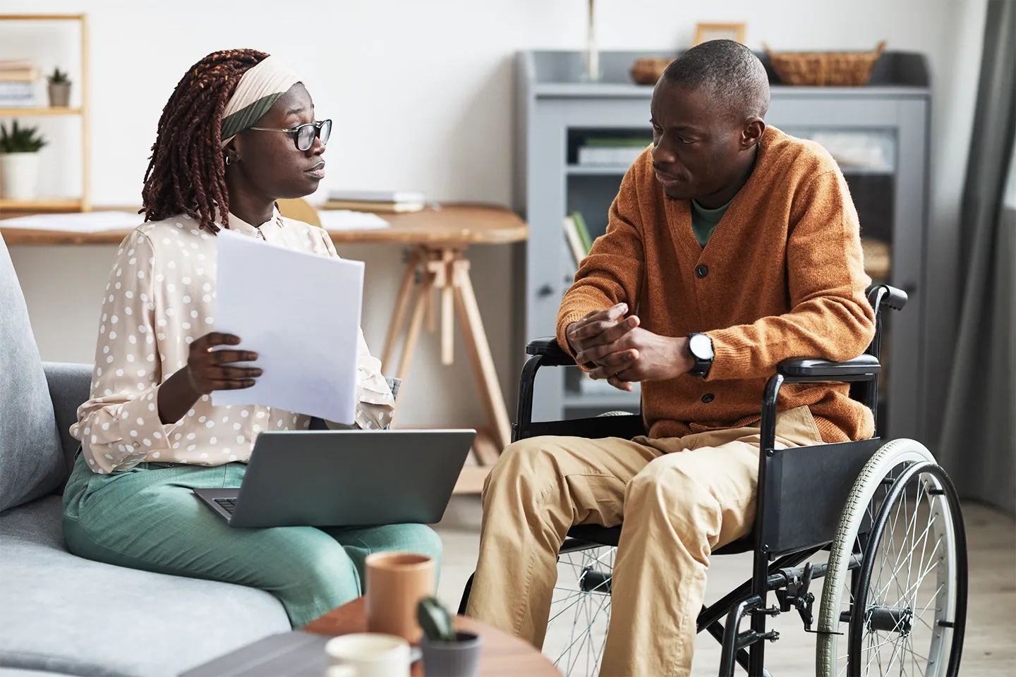 photo of Black woman with laptop speaking to a Black man sitting in a wheelchair in his home