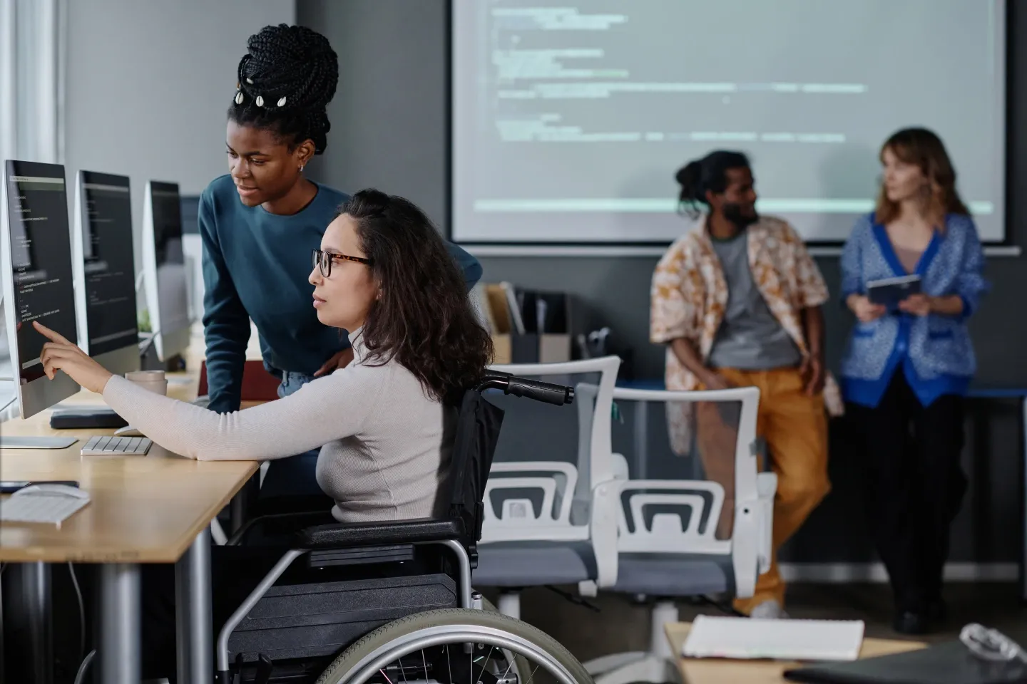 Woman in a wheelchair is sitting at her desk on the computer, pointing to the screen. There is another woman beside her, watching.