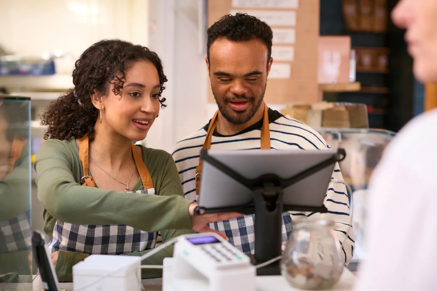 Woman working in food shop training a young man with down syndrome how to use checkout.