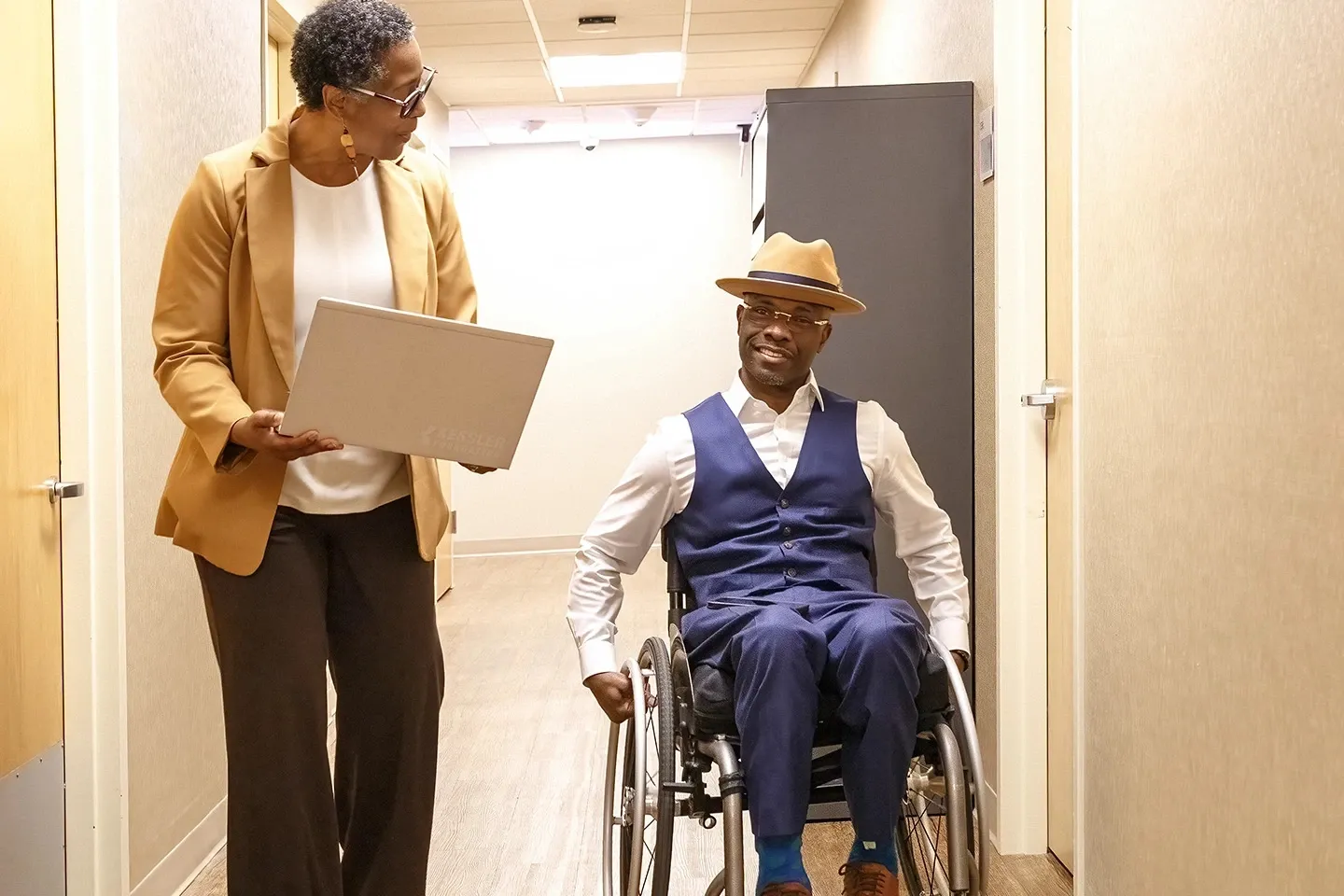 A woman and a man in a wheelchair are heading down a hallway.
