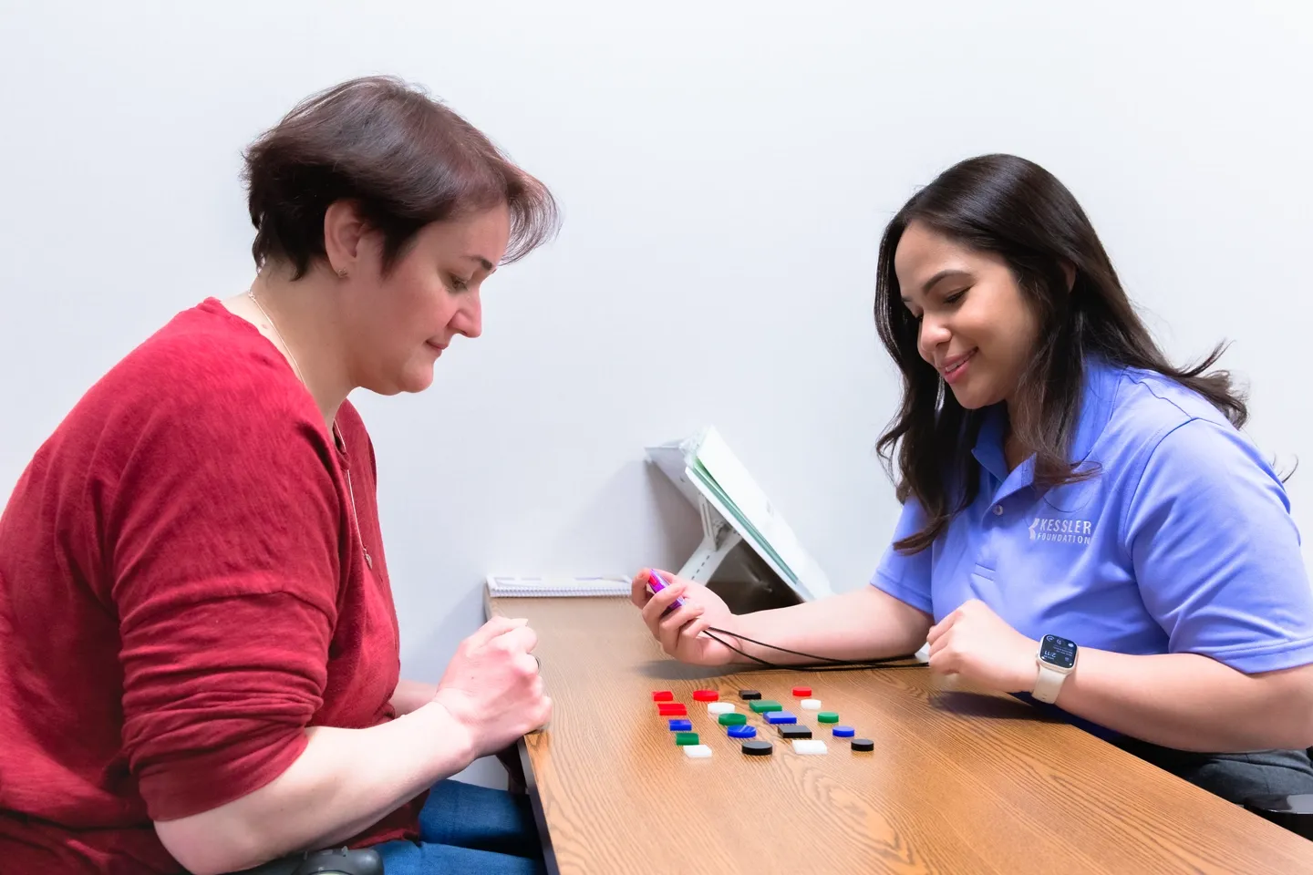 Female test subject wearing a bright red t-shirt sits across the table from a female clinician in blue T-shirt holding a stopwatch with multicolored, differently shaped disks between them.