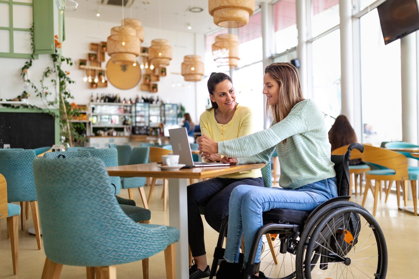 Two women sitting at a wooden table in a bright, modern cafe, engaged in conversation. One woman is in a wheelchair and pointing at a laptop on the table.