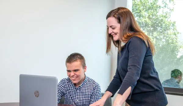 Two individuals collaborating at a desk with an open laptop in a bright office setting, one seated and one standing, reviewing information on the screen 