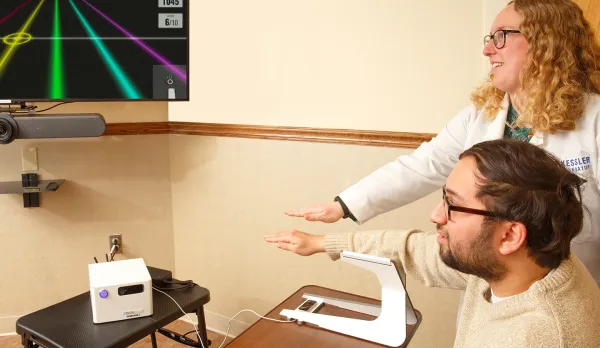 Female clinician shows male stroke participant performing how to perform rehabilitative hand exercises while sitting in front of a screen that shares a gaming element to make the exercises more interactive.