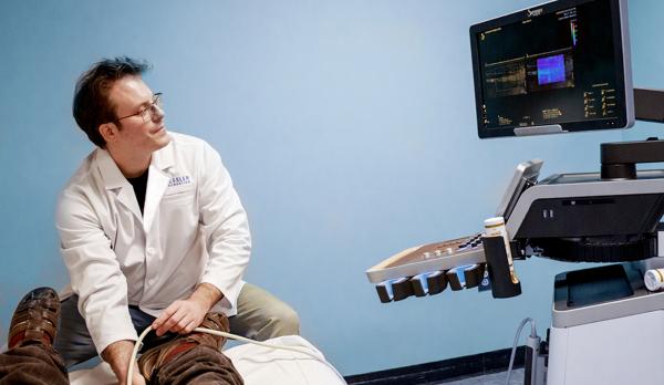 A patient participating in a regenerative rehabilitation session in a clinical setting, seated on an exam table with medical monitoring equipment positioned nearby as part of therapy and assessment.
