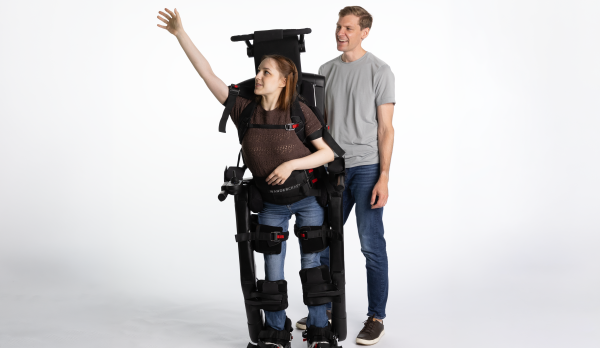 A photo of a woman using Eve™, the first self-balancing personal exoskeleton for eligible wheelchair users. A man in a gray shirt and jeans stands behind her.