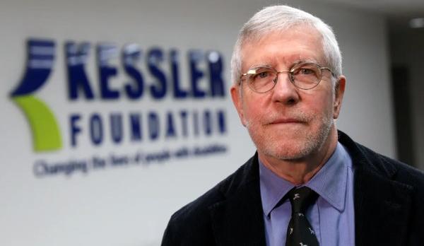 Gray-haired man with glasses, a blue shirt, dark blazer and tie, is pictured in front of a "Kessler Foundation" sign.