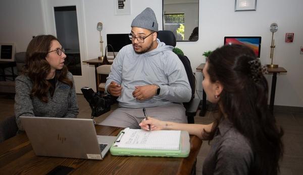 SCI Navigator Program participant Louis Ayala, center, dressed in gray sweats and a gray hat, speaks to two program navigators, Emily DeBel (left), and Samantha "Sam" Rodimer (right).
