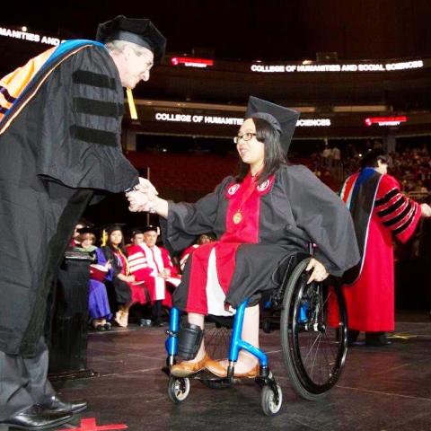 A graduate in a wheelchair on stage, receiving her diploma and shaking hands with a man