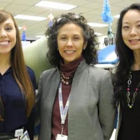 Three women, left brow hair blue blouse, center curly hair grey jacket, right dark hair wearing a white jacket.