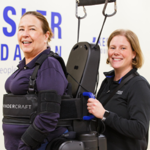 A woman is standing in an exoskeleton with a physical therapist assisting her from behind.
