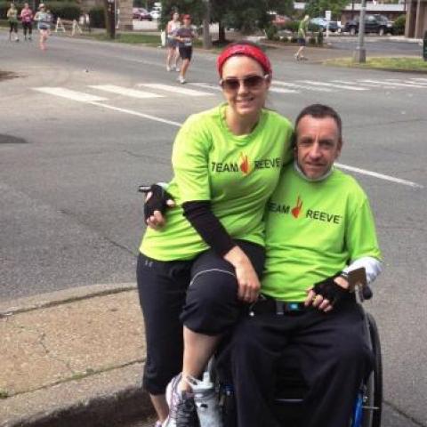 woman next to a man in a wheelchair on the streets, both wearing green t-shirts. 