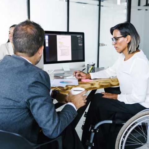 A man in a suit and a woman in a wheelchair are sitting at a desk in an office working together.