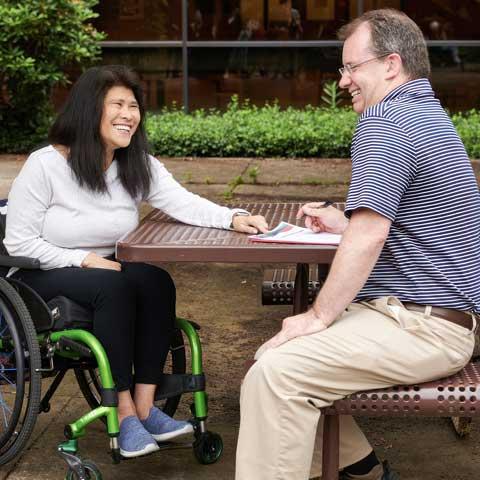 A woman in a wheelchair is sitting at a picnic table next to a man. They are both smiling.