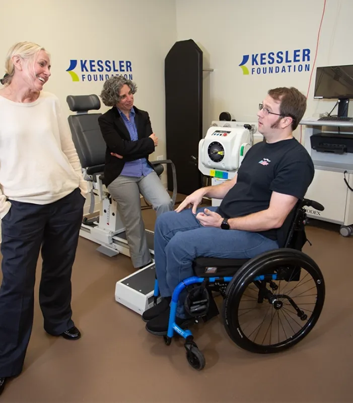 Three individuals in a rehabilitation research lab at Kessler Foundation, including one person using a wheelchair, engaged in discussion near advanced rehabilitation equipment and monitors. 