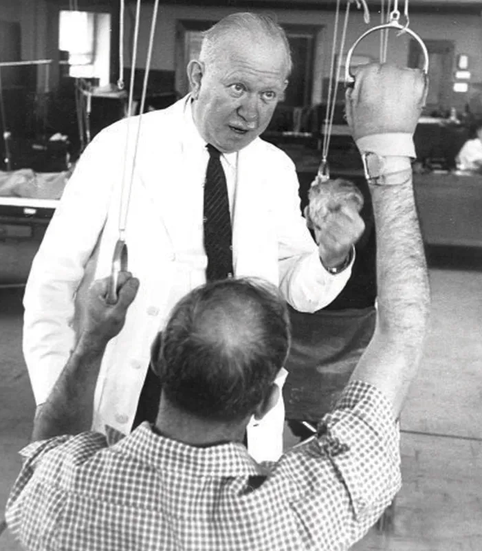 Historic black-and-white photo of a rehabilitation session showing an individual using arm pulley exercise equipment while a clinician in a white coat provides guidance.