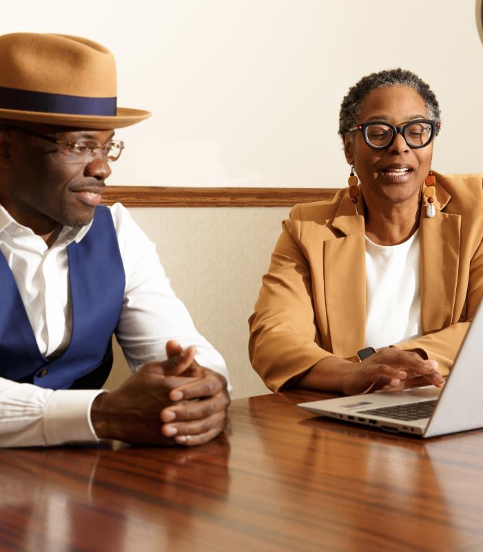 Two people sit side by side at a table, looking at a laptop together. One person gestures while listening, and the other speaks while using the computer, suggesting a collaborative discussion in an indoor office setting.