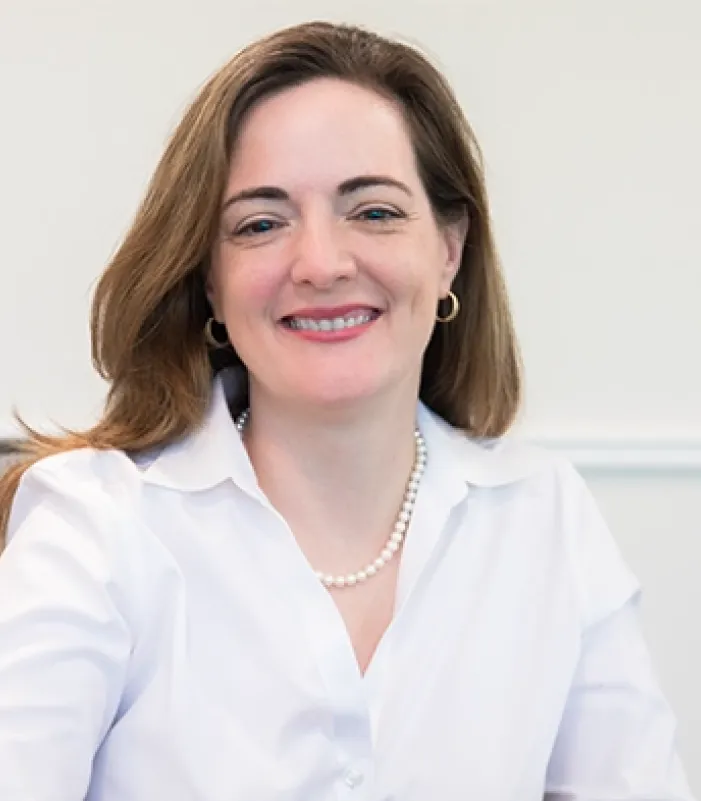 Individual seated in an office chair wearing a white button-down shirt and a pearl necklace, with a light-colored wall and trim in the background. 