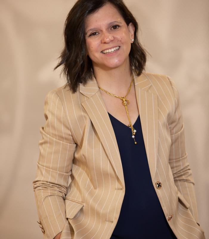 Person wearing a beige pinstripe blazer over a dark navy top, accessorized with a gold necklace, standing with hands in pockets against a neutral background.
