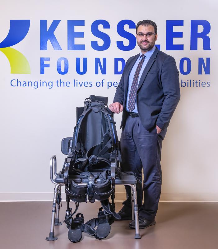 Person wearing a dark suit and tie standing next to a robotic exoskeleton device positioned on a chair, in front of a wall with the Kessler Foundation logo and tagline ‘Changing the lives of people with disabilities