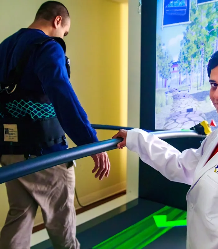 Person wearing a white lab coat standing beside a treadmill, monitoring another individual walking with a safety harness and support bars, in a rehabilitation setting with a virtual reality screen in the background.