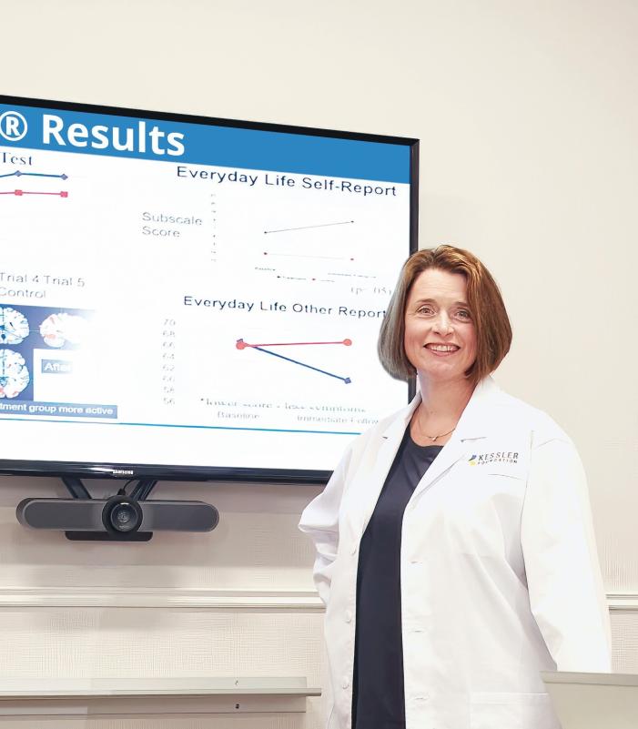 A person wearing a white lab coat stands in front of a wall-mounted monitor displaying research results titled “KF-mSMT® Results.