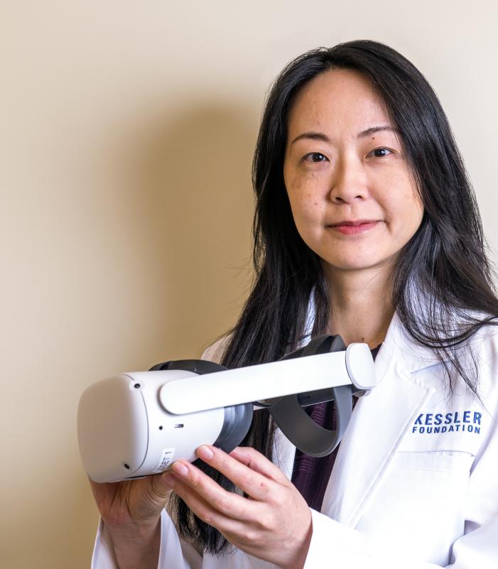 A person wearing a white lab coat with the Kessler Foundation logo is holding a virtual reality headset. The individual is standing against a plain beige background, illustrating the use of VR technology for stroke learning and rehabilitation.