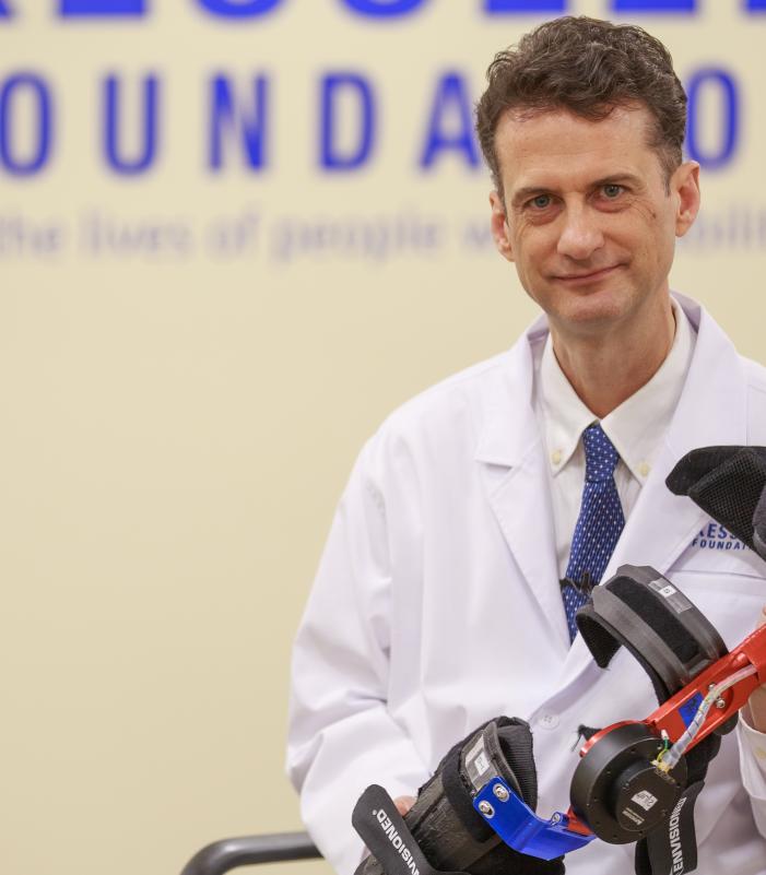 Person wearing a white lab coat and patterned tie holding a black and red orthopedic device, seated in front of a wall with the Kessler Foundation logo and tagline