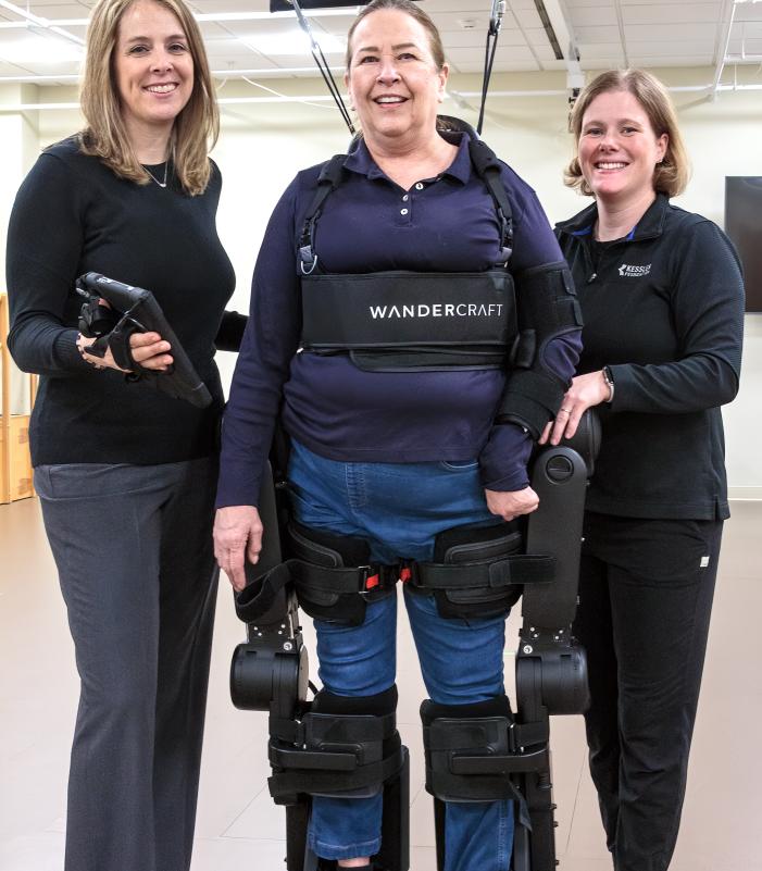 Three individuals in a rehabilitation setting, with one person wearing a robotic exoskeleton labeled ‘Wandercraft’ and supported by an overhead harness system, while two others stand on either side providing assistance and holding a control device