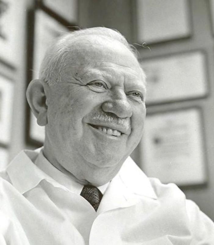 A man smiles in front of a wall of framed art, degrees, and certificates