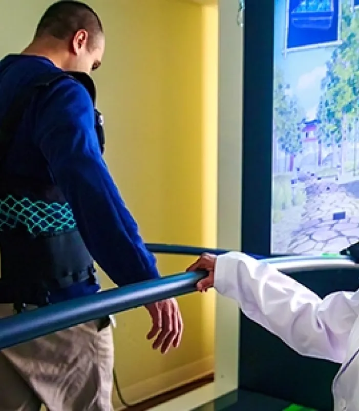 Kiran Karunakaran, PhD wearing a white lab coat standing beside a treadmill, monitoring another individual walking with a safety harness and support bars, in a rehabilitation setting with a virtual reality screen in the background.