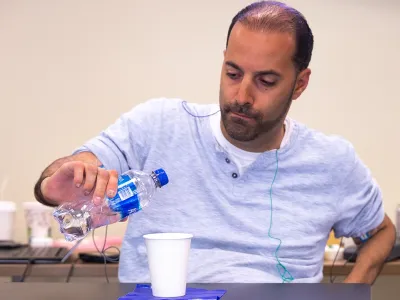Individual seated at a table pouring water from a plastic bottle into a paper cup during a rehabilitation or therapy activity, wearing a light gray shirt with wires attached for monitoring. 