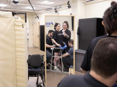 A woman stands assisted during a rehab setting