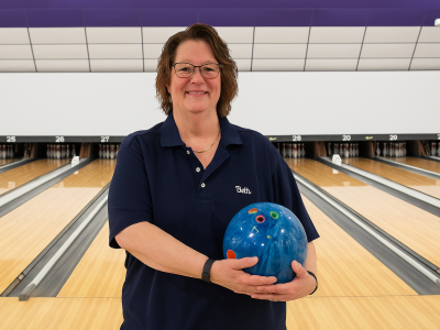 Person standing at a bowling lane holding a blue bowling ball, with polished lanes and pins visible in the background.