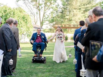 A father using a power wheelchair escorts his daughter down the aisle at her outdoor wedding, as guests stand on either side in a garden setting surrounded by trees and greenery.