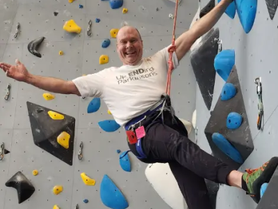 Person wearing a “Up Ending Parkinson’s” T-shirt climbs an indoor rock wall, secured by a rope and harness, stretching one arm outward while balancing on blue and yellow holds against a gray wall.