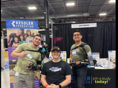 Three adults stand behind a participant using a wheelchair at a Kessler Foundation exhibit booth, with two individuals wearing robotic exoskeleton devices, demonstrating rehabilitation technology in a conference setting.