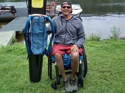 A wheelchair user sits on grass near a lake, posing beside upright  outdoor gear, with calm water, boats, and a green shoreline in the background.
