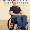 Individual using a wheelchair seated in front of a Kessler Foundation wall with the tagline ‘Changing the lives of people with disabilities,’ wearing a black shirt and blue pants. 