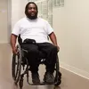 Individual using a wheelchair moving through a hallway with light-colored walls and framed displays, wearing a white shirt and dark pants.